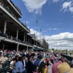Crowded grandstand at a sunny outdoor horse race, spectators seated along the railing. ️