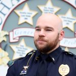 Portrait of a bald police officer in a dark uniform with a gold badge and name tag 'E. Kruse', standing in front of a decorative backdrop with stars and signage. (Informative)