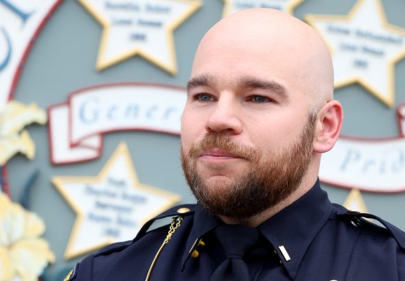 Portrait of a bald police officer in a dark uniform with a gold badge and name tag 'E. Kruse', standing in front of a decorative backdrop with stars and signage. (Informative)