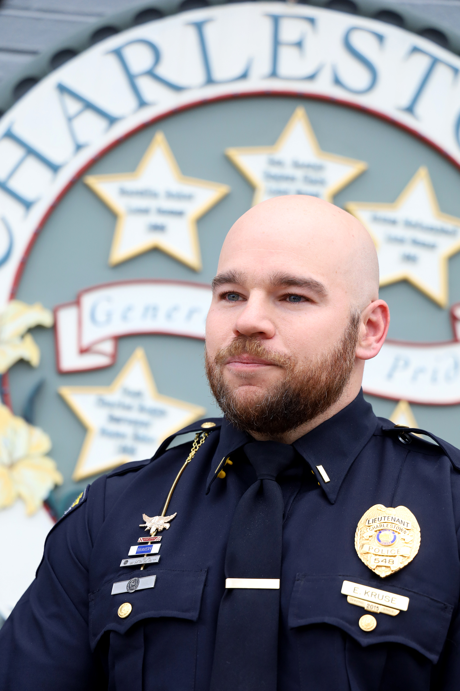 Portrait of a bald police officer in a dark uniform with a gold badge and name tag 'E. Kruse', standing in front of a decorative backdrop with stars and signage. (Informative)