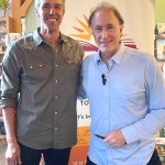 Two men smile for a photo indoors, standing side by side in front of a banner with a sunburst design.