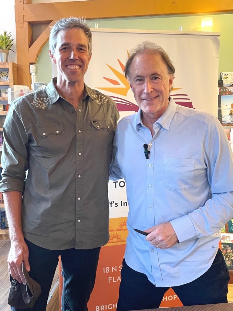 Two men smile for a photo indoors, standing side by side in front of a banner with a sunburst design.