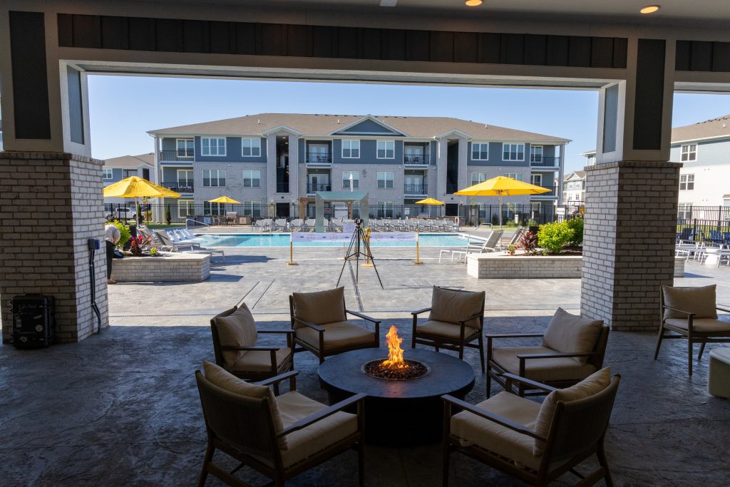 Arlington Farms RC 572026 FS-127 - Extol Media Outdoor lounge around a circular fire pit with cushioned chairs, facing a swimming pool and yellow umbrellas in a residential complex.