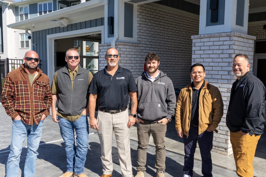 Arlington Farms RC 572026 FS-19 - Extol Media Seven men stand side by side outside a brick building, wearing casual workwear and sunglasses, posing for a group photo.