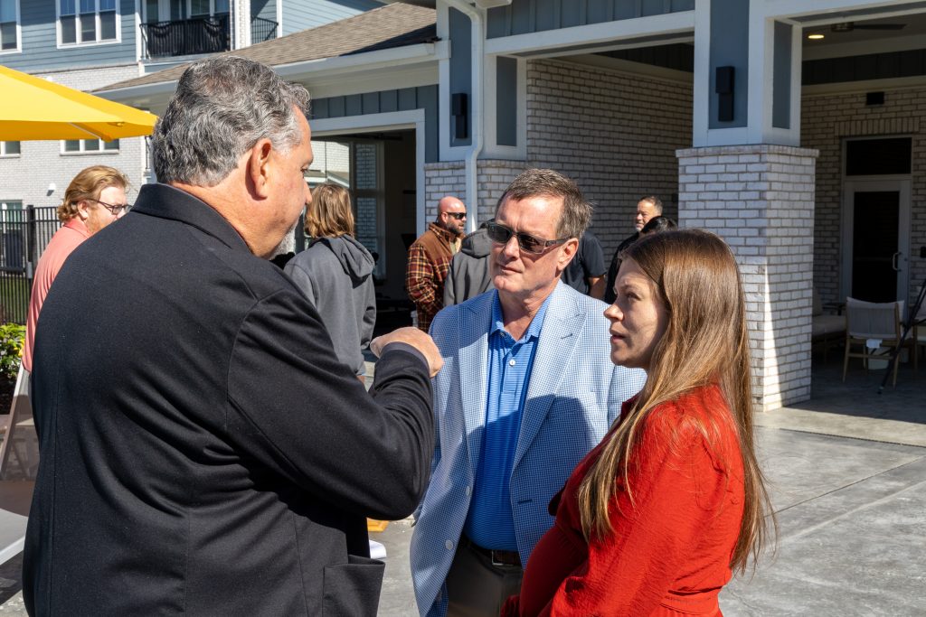 Arlington Farms RC 572026 FS-27 - Extol Media Group of people conversing outdoors near a building with brick columns at a sunny event.