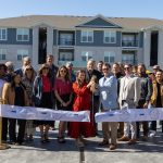 Diverse group of adults at a ribbon-cutting ceremony outside a modern apartment complex.