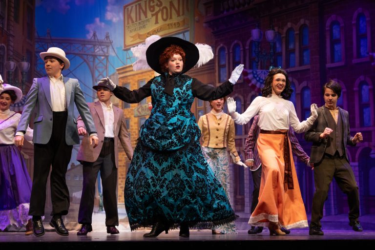 Theatrical performance with performers in 19th-century costumes dancing on a stage; center features a woman in a teal patterned dress and large black hat with white feathers.