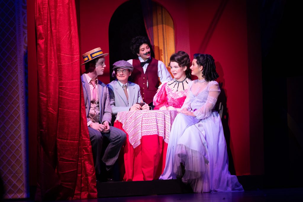 Five performers in vintage costumes gathered around a lace-covered table on a red-curtained stage, lit in pinkish light.