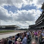 Crowd of spectators wearing hats seated in a multi-tiered grandstand at a racetrack, with a dirt track and fencing in the foreground under a partly cloudy sky.