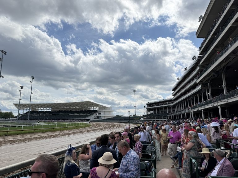 Crowd of spectators wearing hats seated in a multi-tiered grandstand at a racetrack, with a dirt track and fencing in the foreground under a partly cloudy sky.