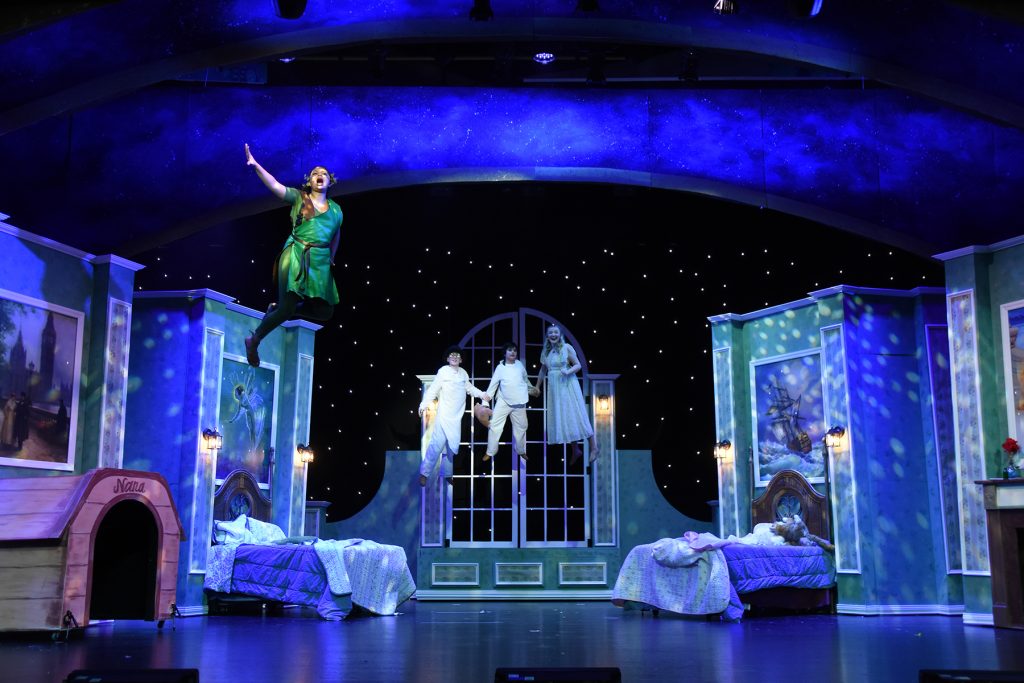 Actor in a green costume leaps mid-air on a theater stage, reaching upward with other performers in white on a balcony behind. Termed a fantastical scene amid blue, starry backdrop and colorful set pieces.