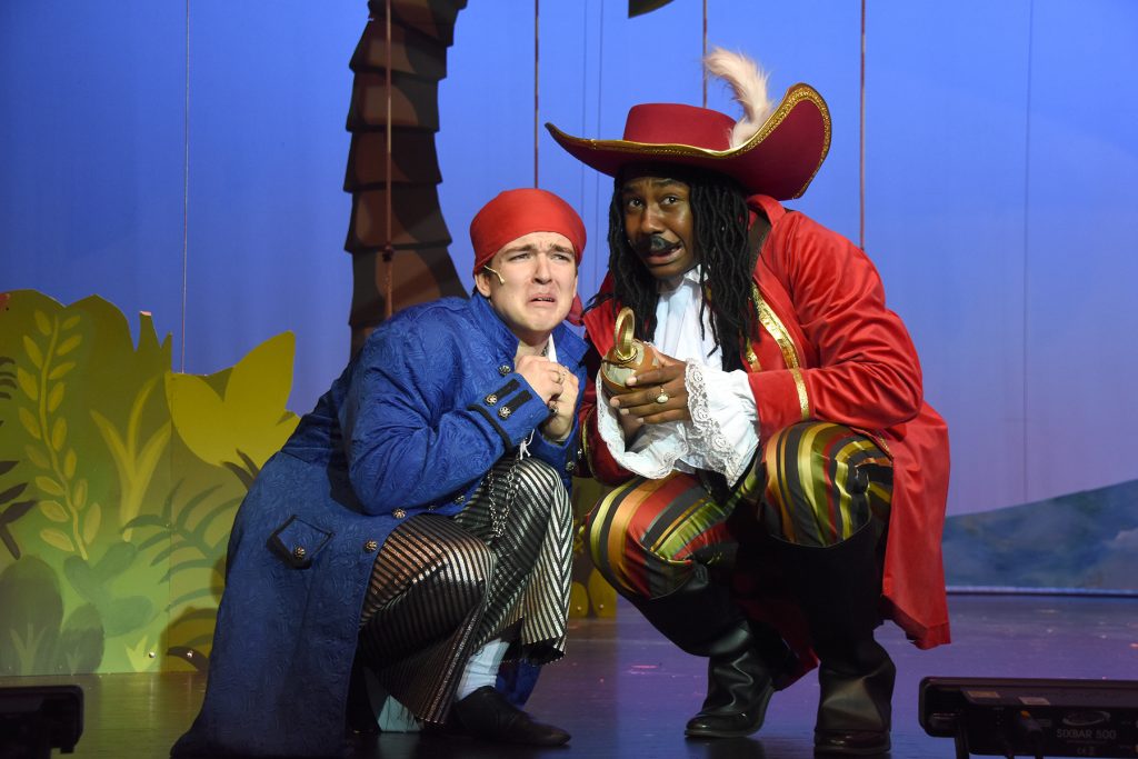 Two costumed pirates crouch on a theater stage, one in a blue coat and red bandana, the other in a red coat and oversized hat, clutching a small goblet.