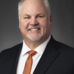 Portrait of a smiling man in a dark suit and orange patterned tie against a grey studio backdrop.