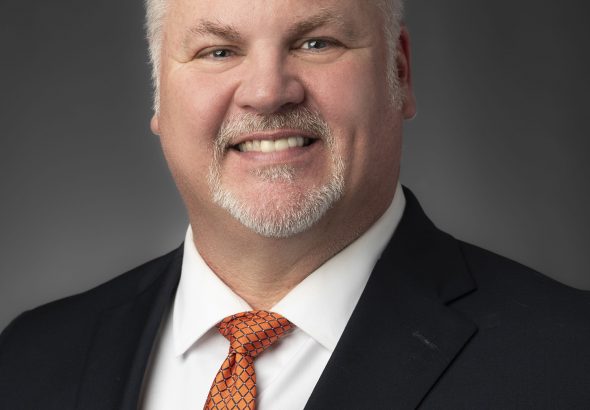 Portrait of a smiling man in a dark suit and orange patterned tie against a grey studio backdrop.