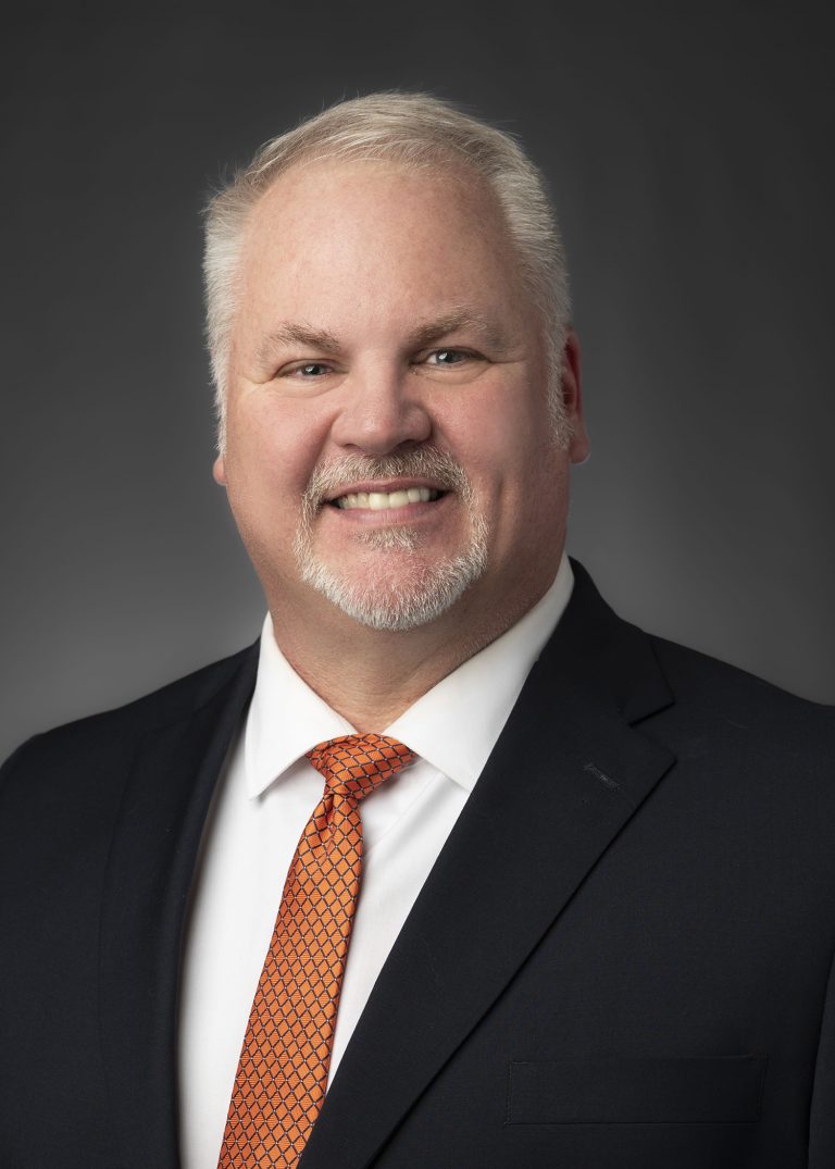 Portrait of a smiling man in a dark suit and orange patterned tie against a grey studio backdrop.
