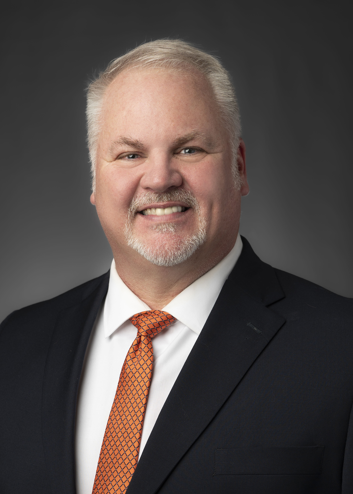 Portrait of a smiling man in a dark suit and orange patterned tie against a grey studio backdrop.
