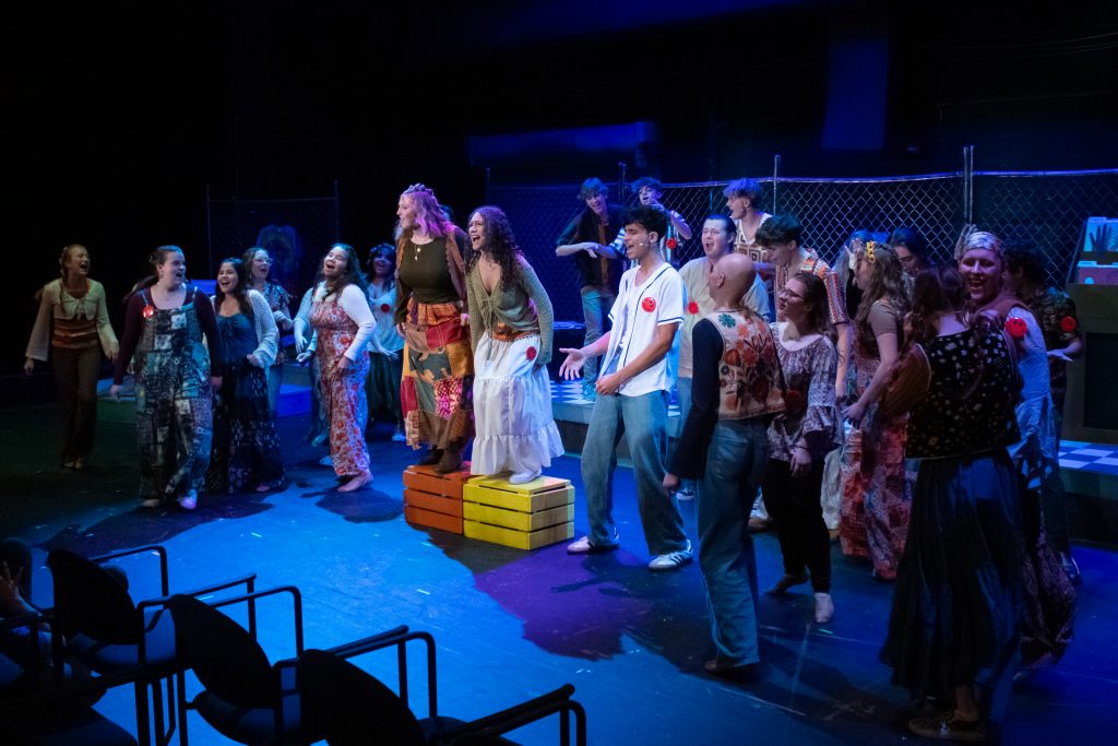 A large group of performers in colorful patchwork outfits sings on a dimly lit stage, with a person standing on stacked blocks at center.