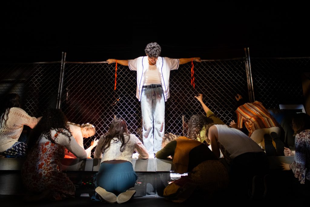 Performer in a white jacket on a dark stage with arms outstretched behind a chain-link fence; audience kneels and reaches toward him.