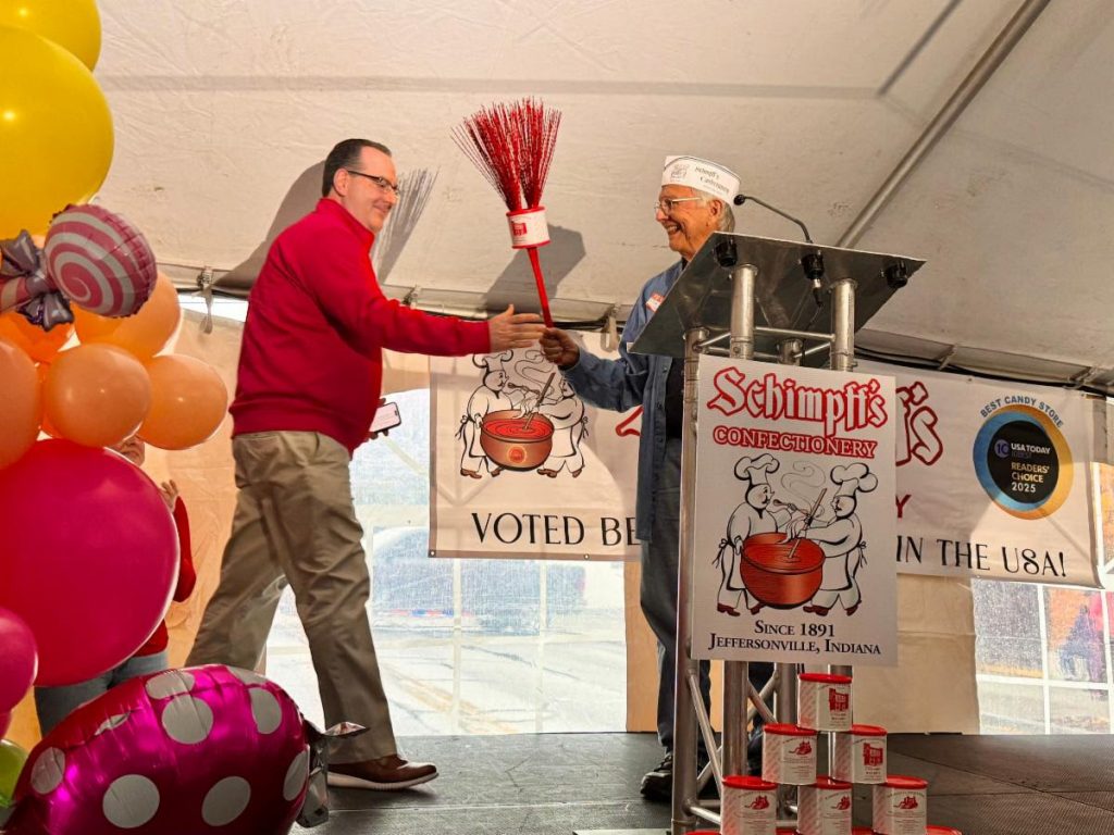 Two men on a stage exchange a red broom while smiling, with candy-themed banners and balloons nearby.