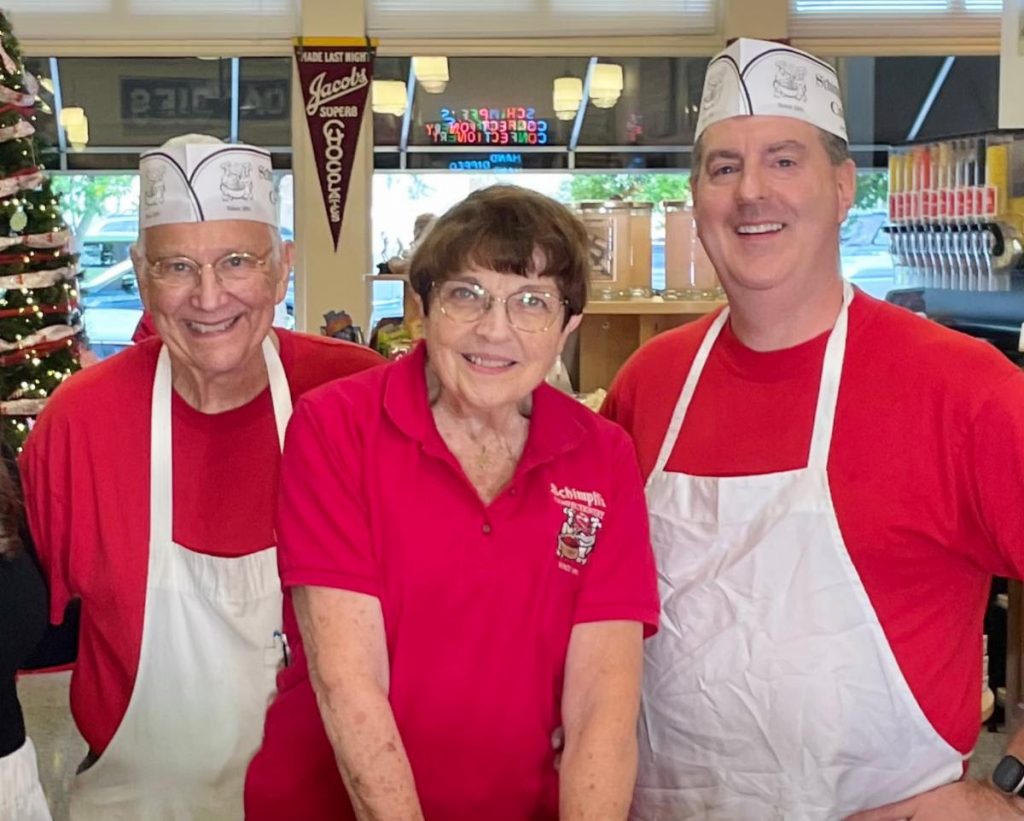 Three smiling volunteers in red shirts and white aprons stand together in a cafe, with a Christmas tree visible on the left.