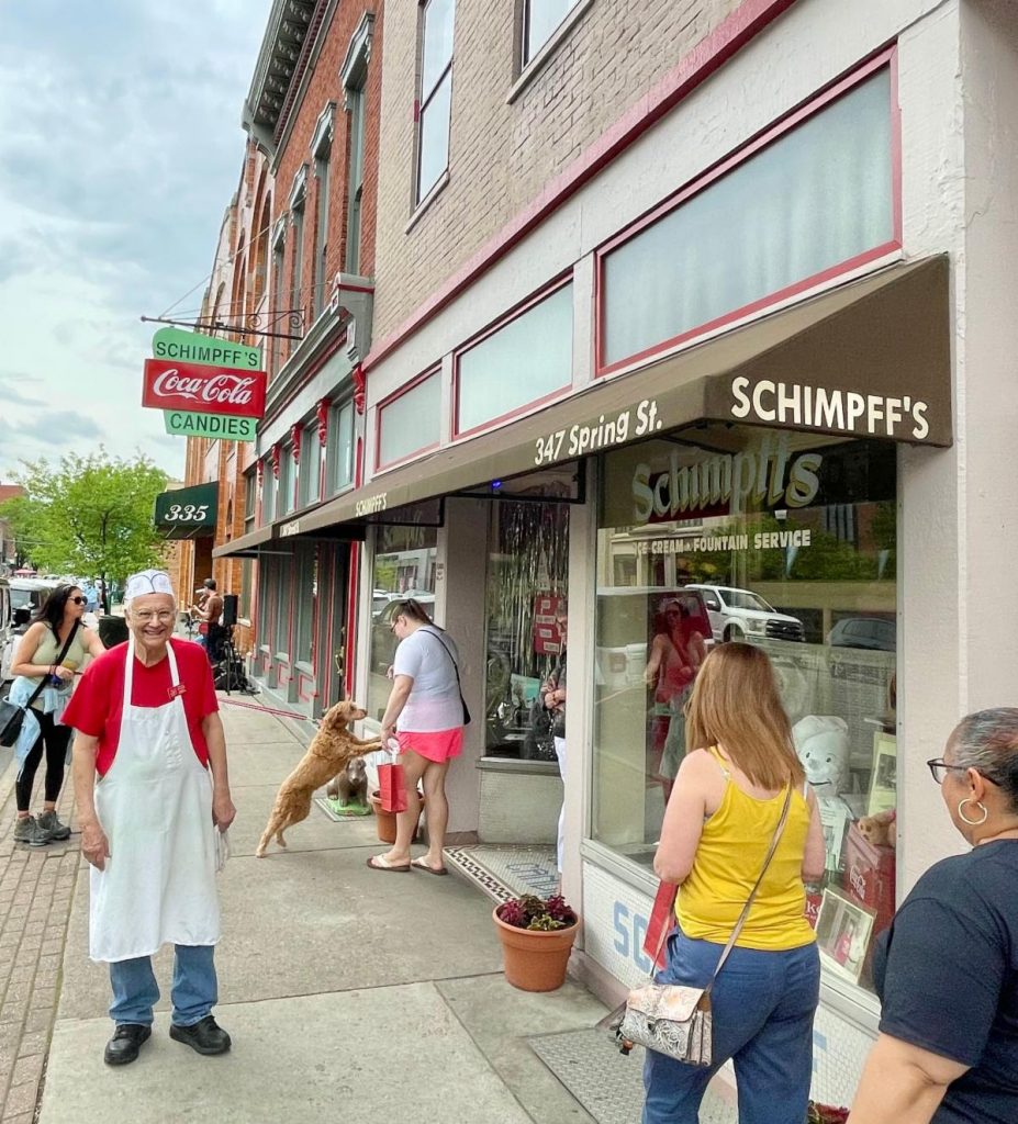 Street scene outside Schimpff's Candies with pedestrians; an elderly man in a red shirt and white apron on the sidewalk while a dog leaps toward a passerby nearby.