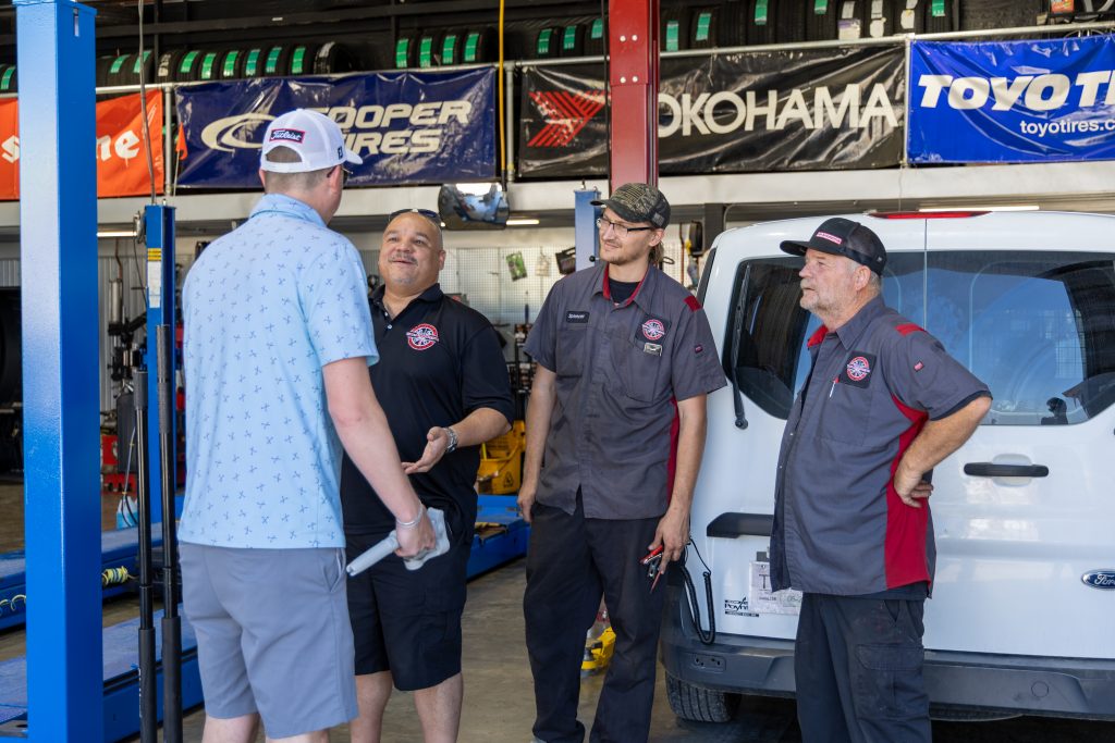 Three men in a garage auto shop talk beside a white van, banners for tire brands in the background.