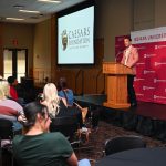 Speaker at a wooden podium delivering a talk on a stage; projection screen shows Caesars Foundation logo beside an Indiana University Southeast backdrop; audience seated.