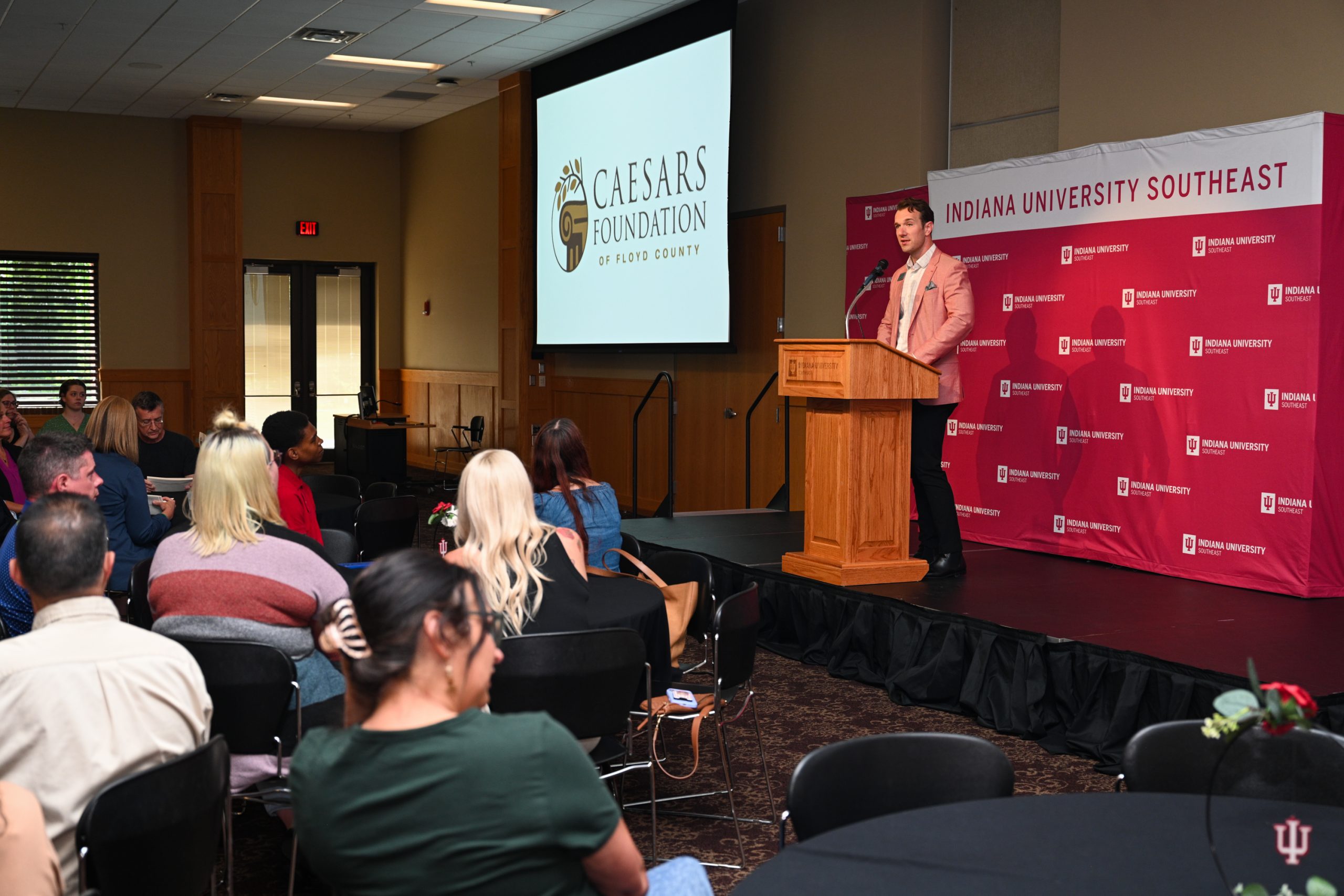 Speaker at a wooden podium delivering a talk on a stage; projection screen shows Caesars Foundation logo beside an Indiana University Southeast backdrop; audience seated.