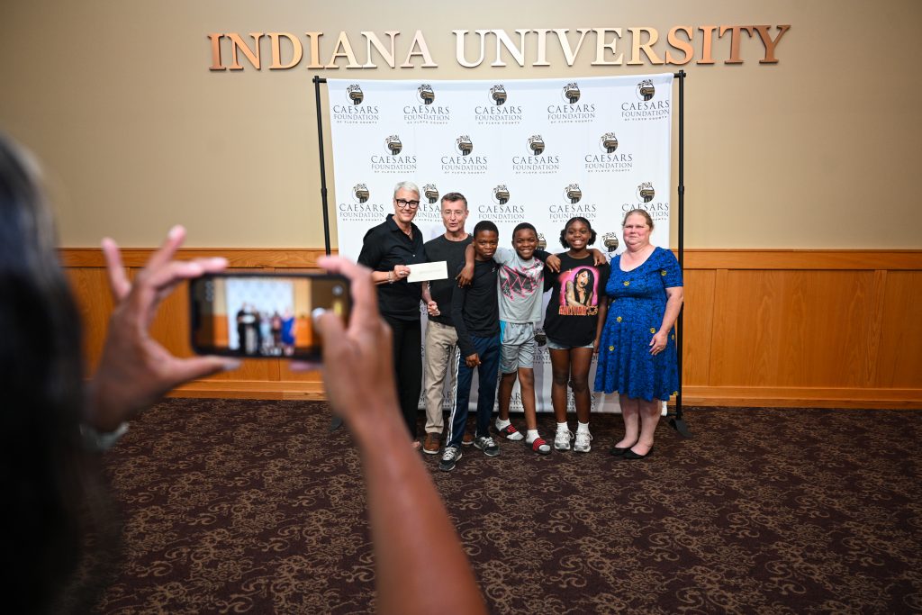 Caesar Grant Awards 06-24-26-62 - Extol Media Group of six people posing for a photo in front of a Caesars Foundation backdrop inside a room, with a person in the foreground holding up a phone to capture the moment.
