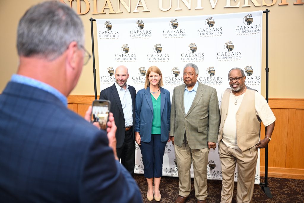 Caesar Grant Awards 06-24-26-72 - Extol Media Five adults pose for a photo in front of a Caesars Foundation backdrop, while a person in the foreground records them with a smartphone.