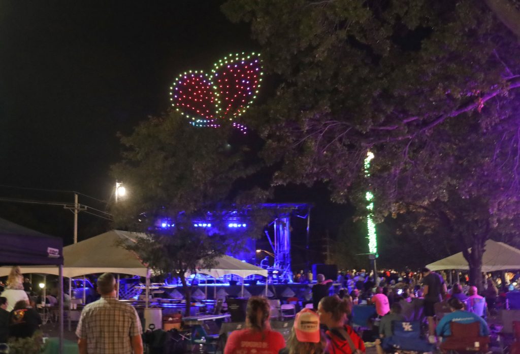 Nighttime festival scene with a large heart-shaped LED light display in red, green, and blue above a crowd.
