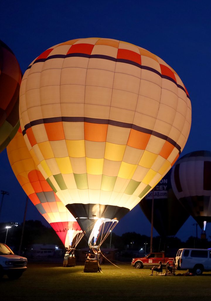 Night scene of a large illuminated hot air balloon about to take off on a grassy field with support vehicles nearby.