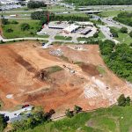 Aerial view of a large construction site with earthwork, excavators, and piles of dirt near roads and buildings.