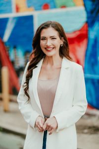 Smiling woman in a white blazer and beige top posing outdoors with a colorful mural in the background.