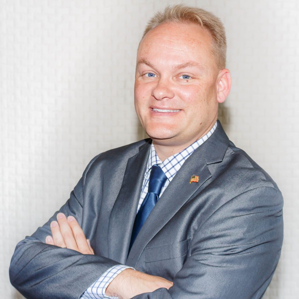 Smiling man in a gray suit with blue tie, arms crossed, wearing an American flag lapel pin.