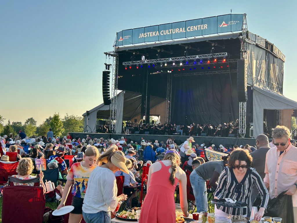 Outdoor music festival with a large stage and banner, crowds seated on lawn chairs enjoying the show at sunset.
