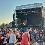 Outdoor music festival with a large stage and banner, crowds seated on lawn chairs enjoying the show at sunset.