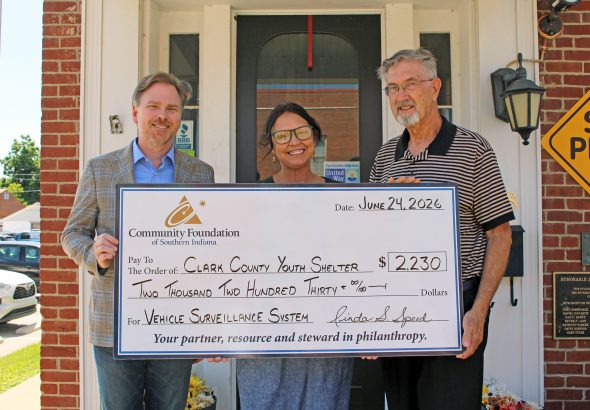 Three adults smile while holding a large donation check outside a brick building, addressed to Clark County Youth Shelter under a Community Foundation of Southern Indiana grant.