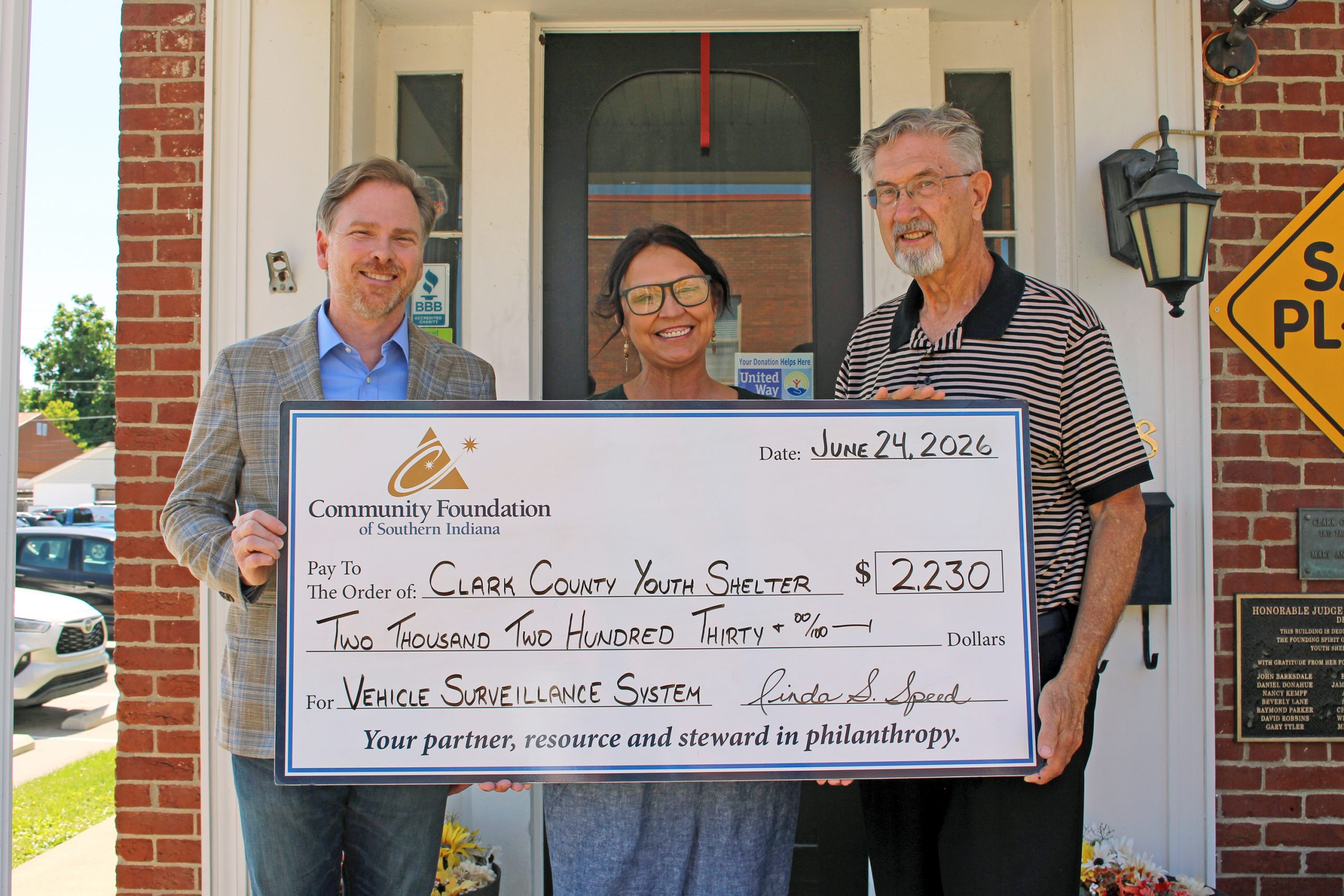 Three adults smile while holding a large donation check outside a brick building, addressed to Clark County Youth Shelter under a Community Foundation of Southern Indiana grant.