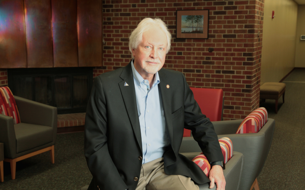 Older man in a blazer and light blue shirt seated in a lounge with a brick wall and fireplace behind him.