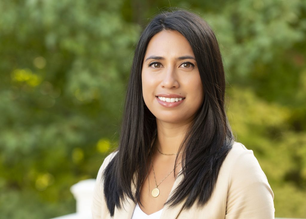 Portrait of a woman with long dark hair, smiling outdoors, wearing a beige blazer and layered gold necklaces.