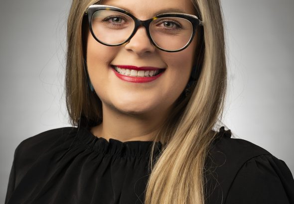 Portrait of a smiling woman with long blonde hair, wearing black-rimmed glasses and a black top against a neutral gray background.