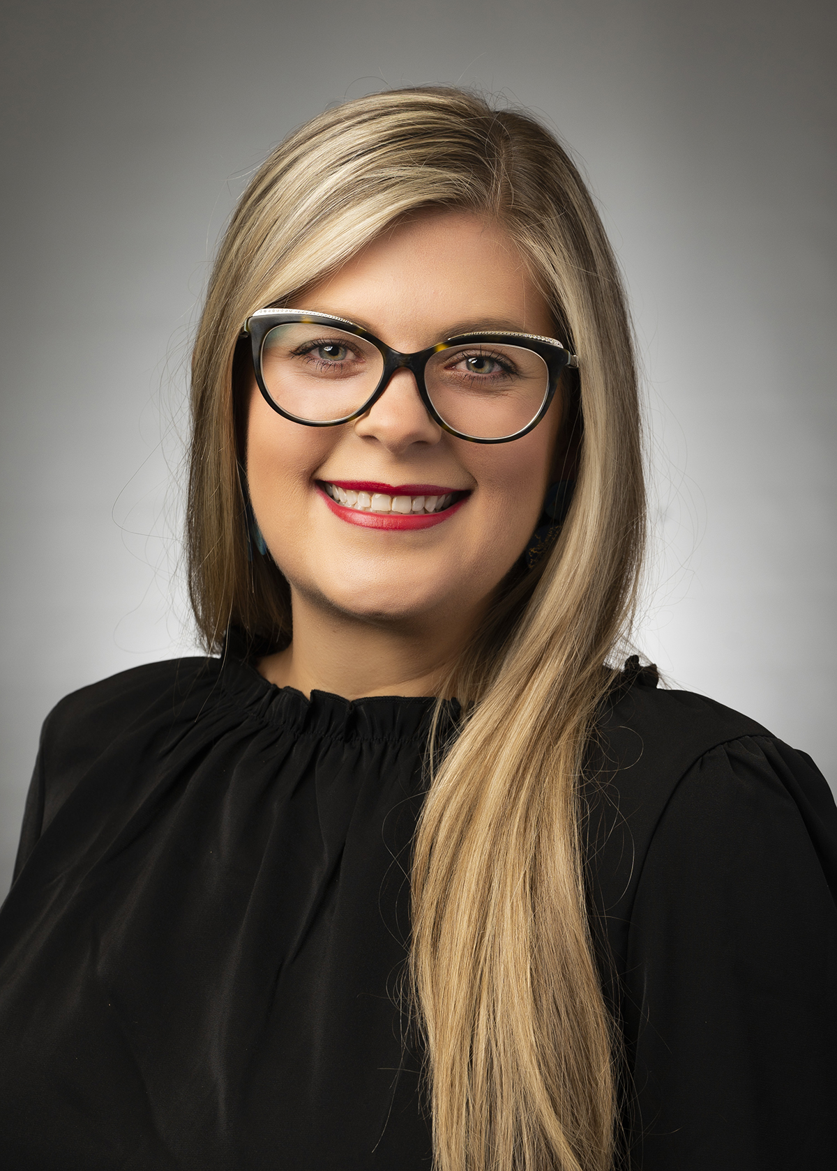 Portrait of a smiling woman with long blonde hair, wearing black-rimmed glasses and a black top against a neutral gray background.